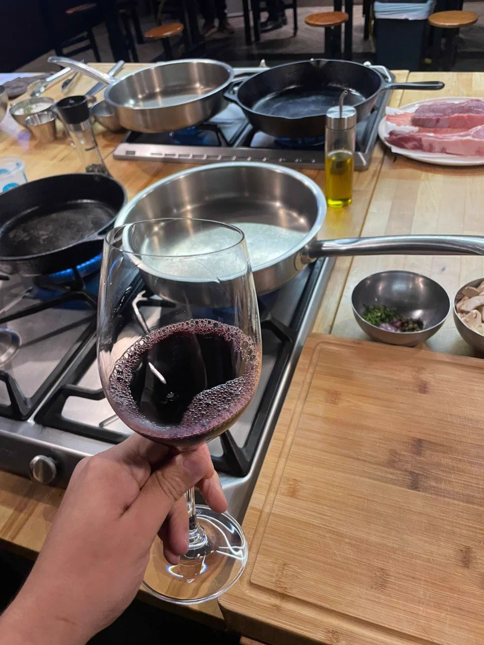 A glass of red wine in front of a cooking station with pans, ingredients, and steak cuts during a culinary experience.